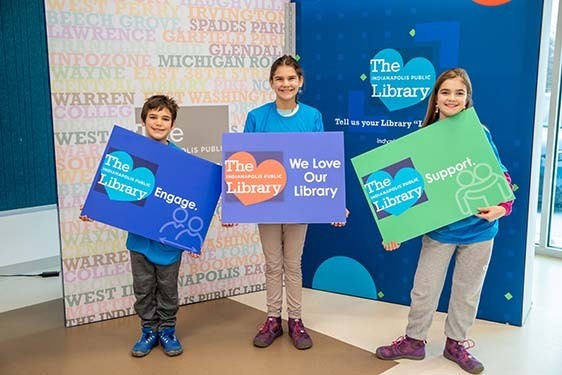 Three smiling kids hold We Love Our Library signs.
