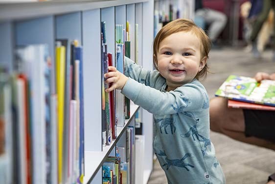 A toddler smiles while looking at all the books on Library shelves.
