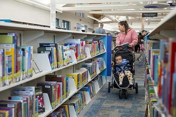 A mother pushing a baby in a stroller looks at books on Library shelves.
